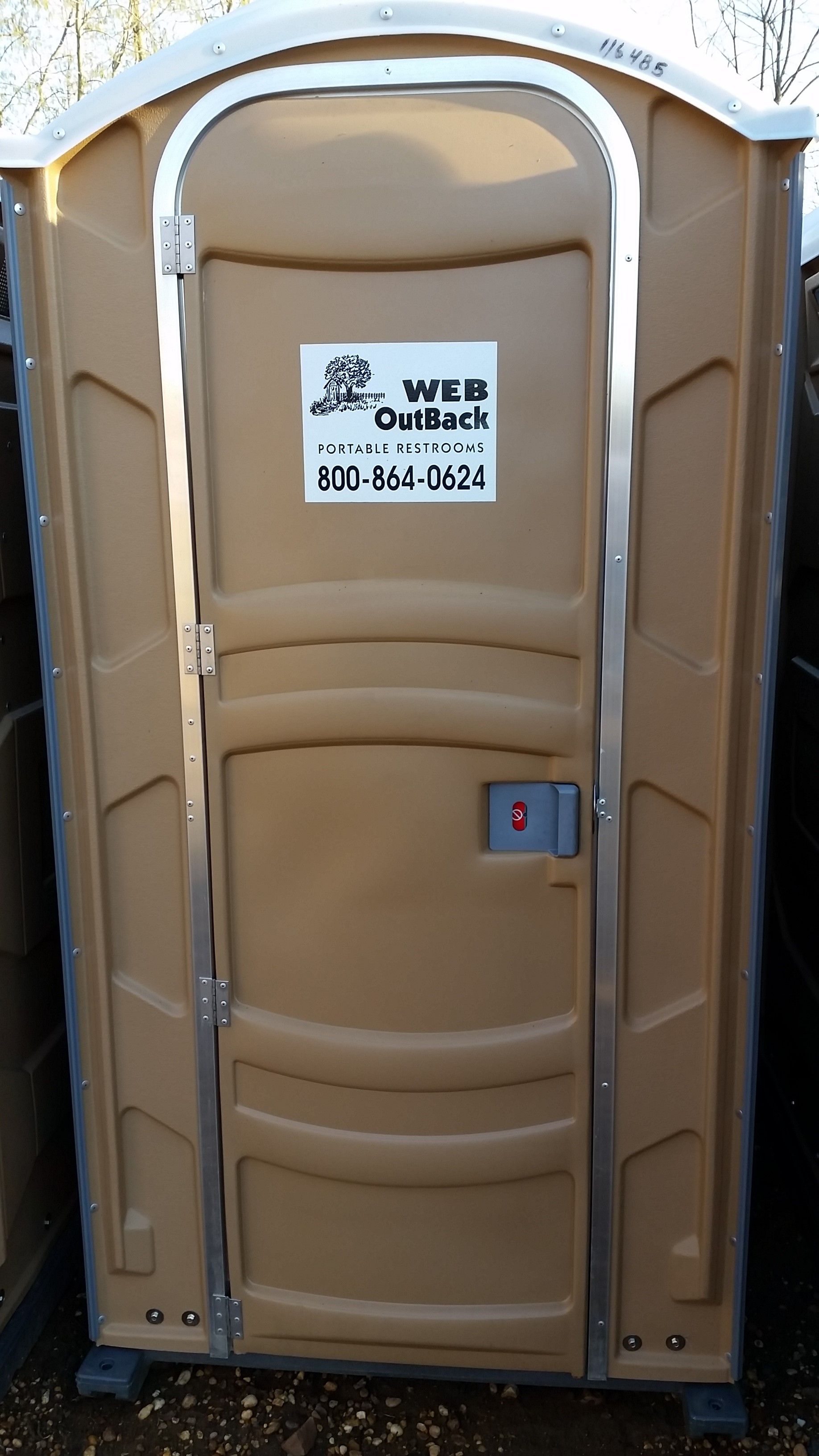 Brown portable toilet with white roof, front door closed, standing outdoors on a dirt surface.
