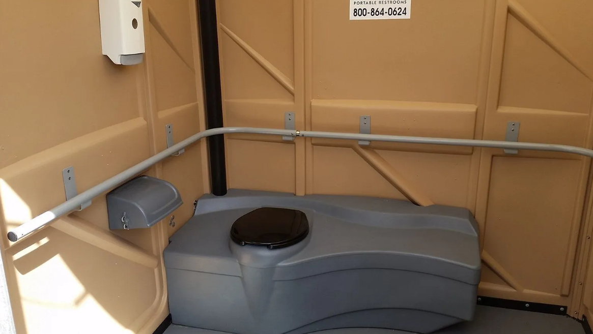 Interior of a portable toilet with a grey toilet, handrail, and soap dispenser.