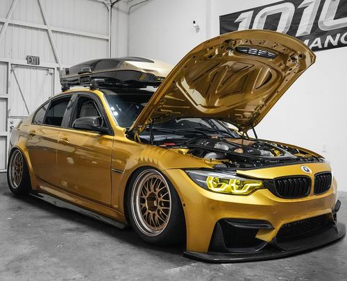 Gold BMW coupe with hood open and black roof box in a garage, yellow headlights on, lowered stance.