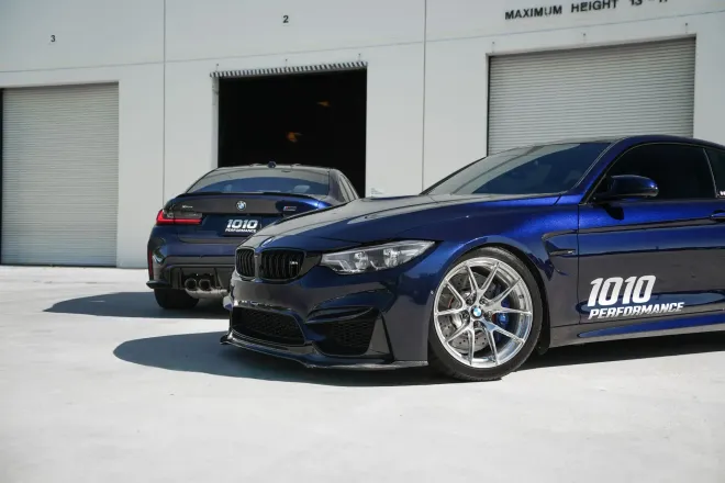 Two dark blue sports cars parked outside garage bays, one with white 1010 decal in front.
