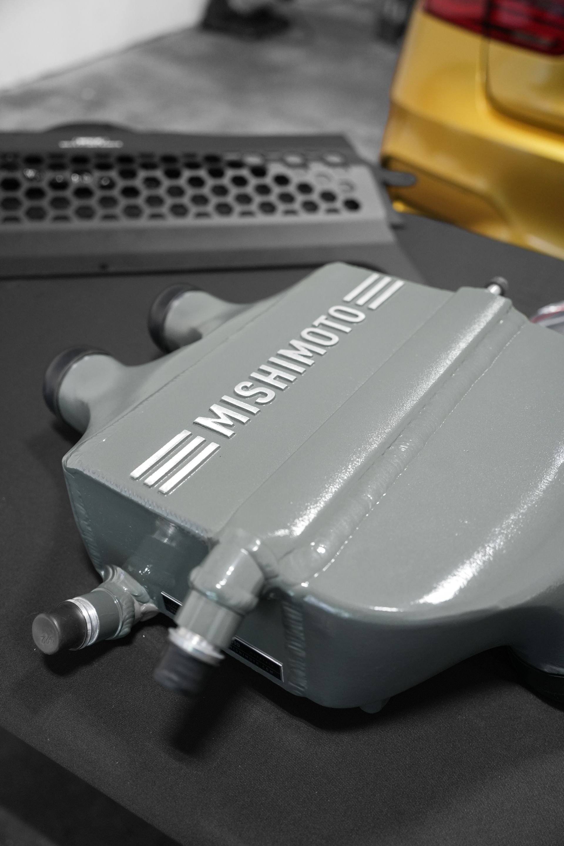 Gray Mishimoto automotive part with silver lettering on a black surface, beside a yellow container.