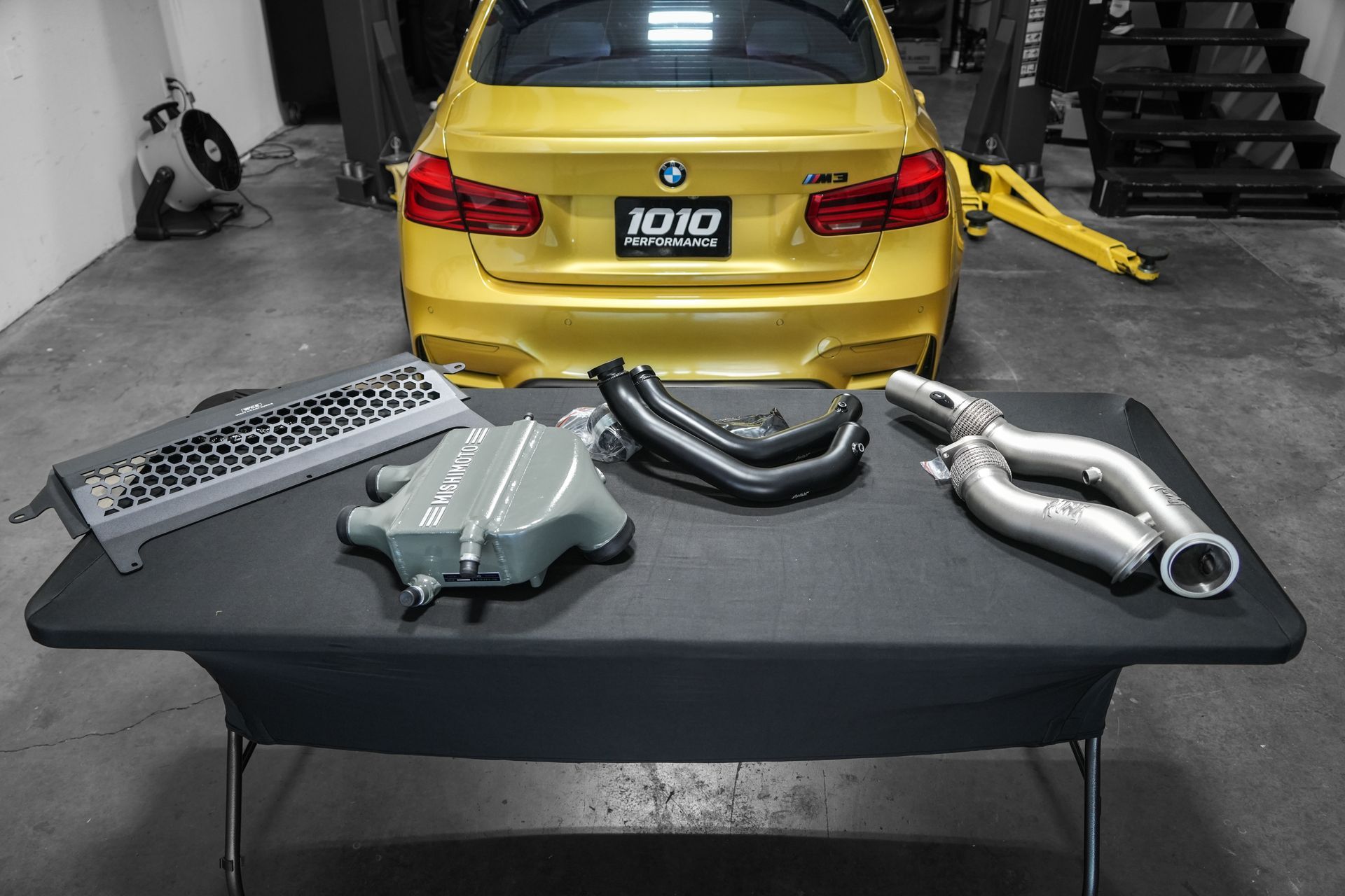 Car parts on a table in a garage, with a yellow car and lift in the background