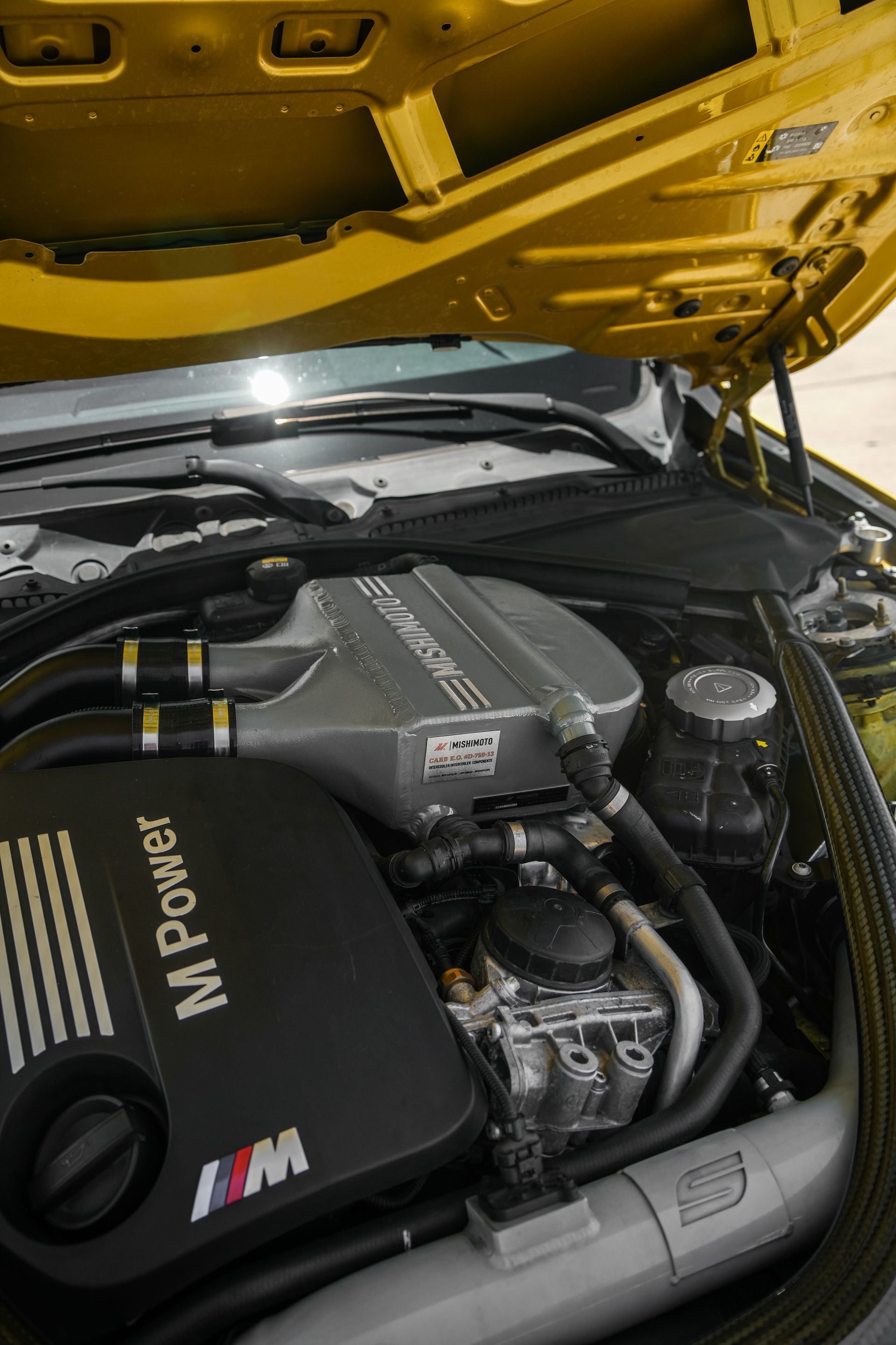 BMW M Power engine bay under a raised yellow hood, showing black performance components and hoses