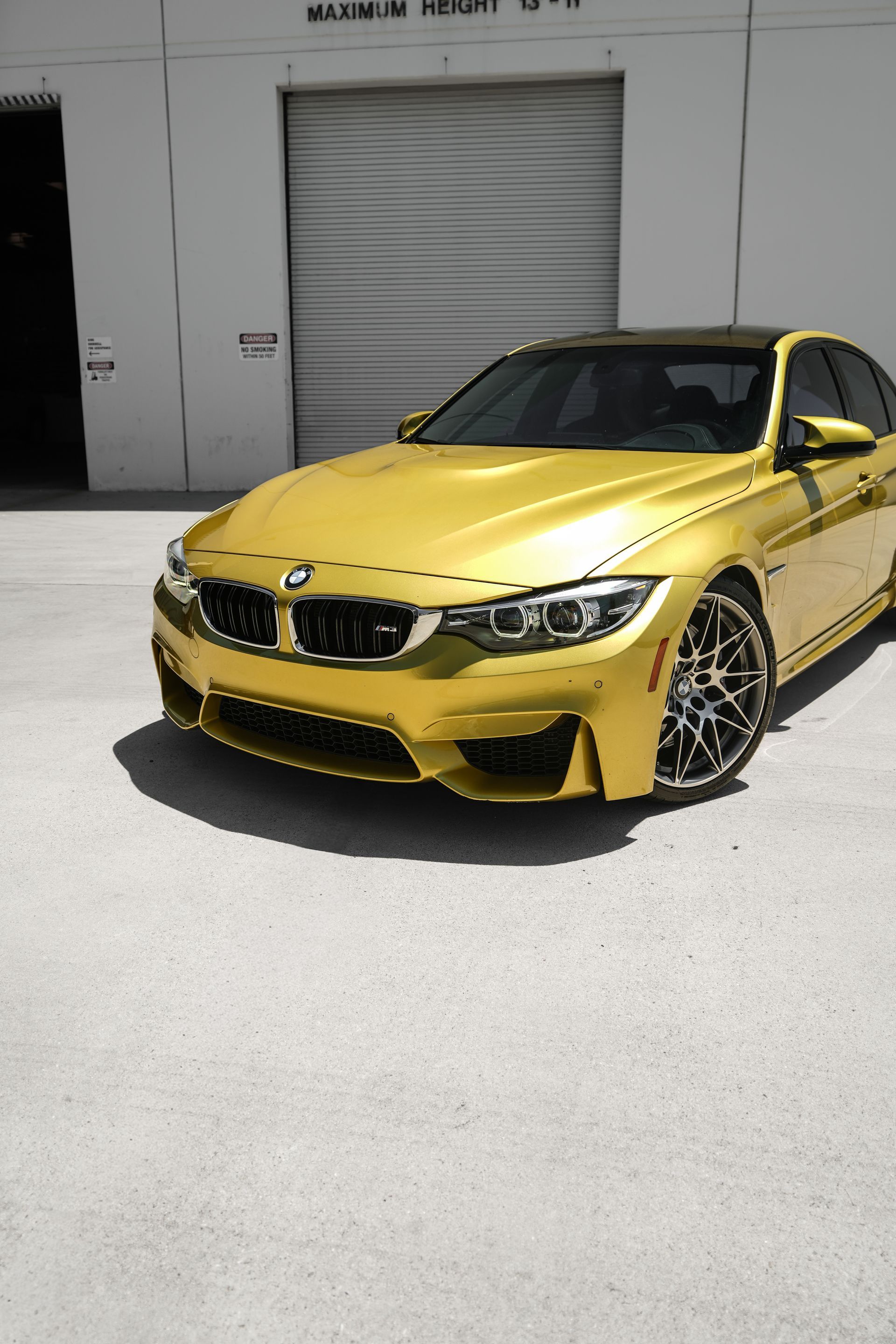 Yellow BMW sedan parked outside a warehouse, front three-quarter view in bright sunlight
