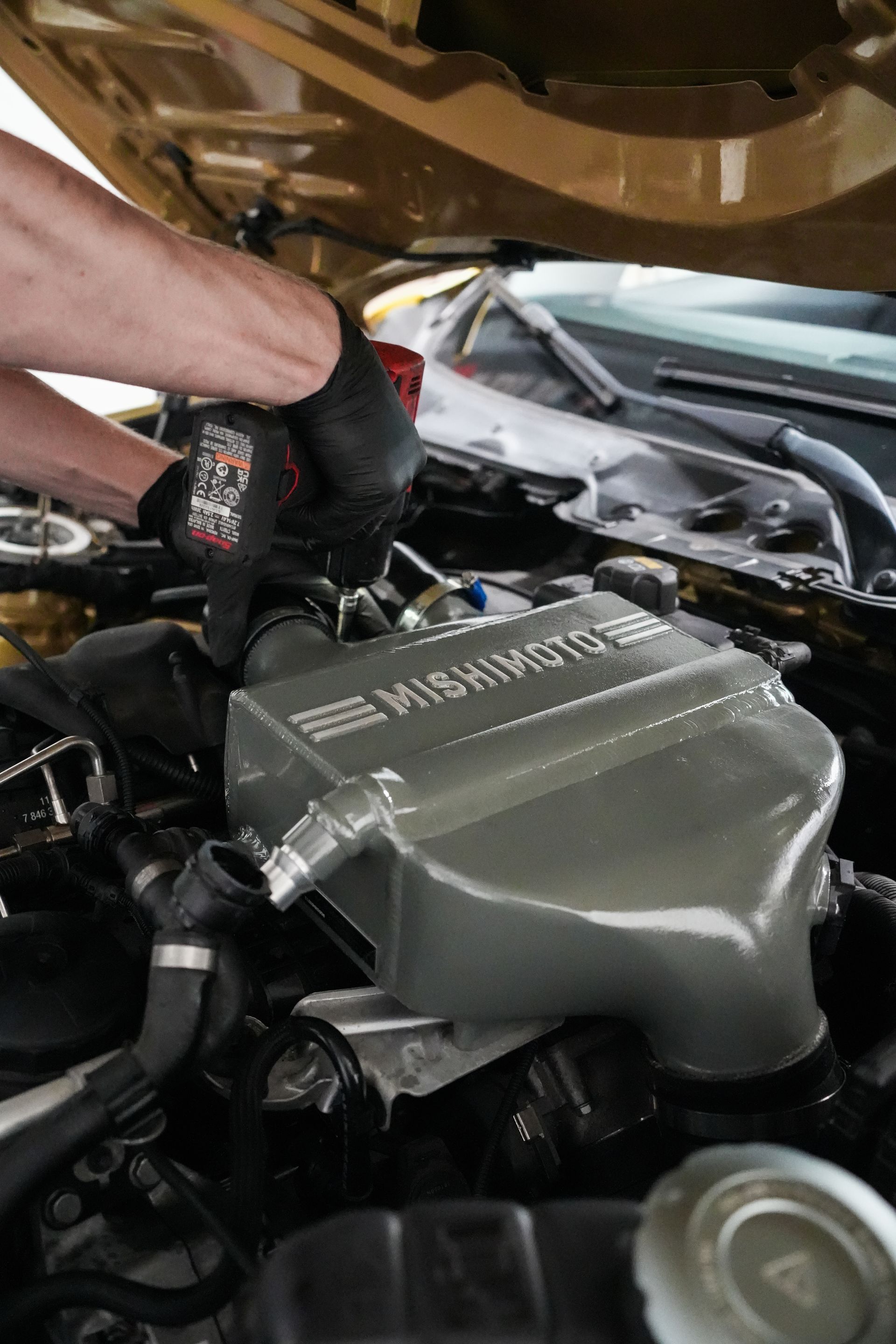Person working under an open car hood with a black glove, adjusting parts near the engine compartment