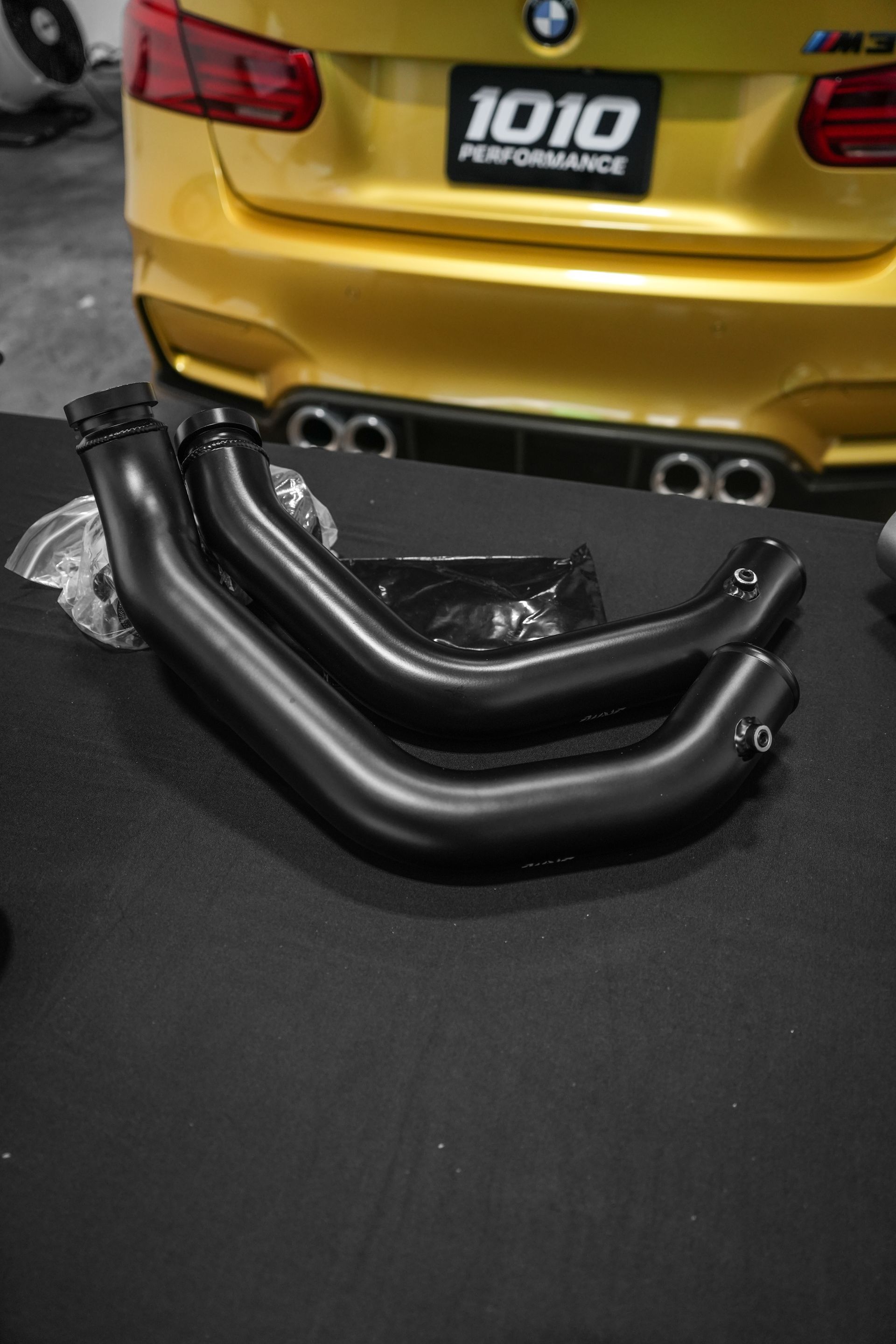 Two black exhaust pipes on a table in front of a gold car rear bumper.