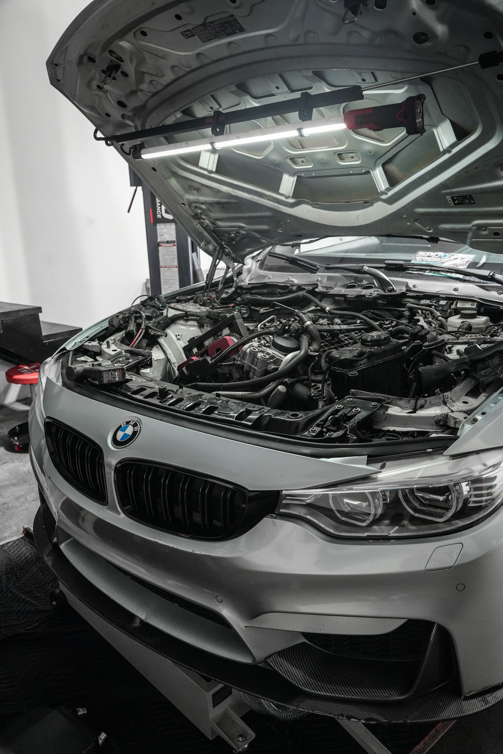 Silver BMW with hood open in a garage, exposing the engine and front damage.