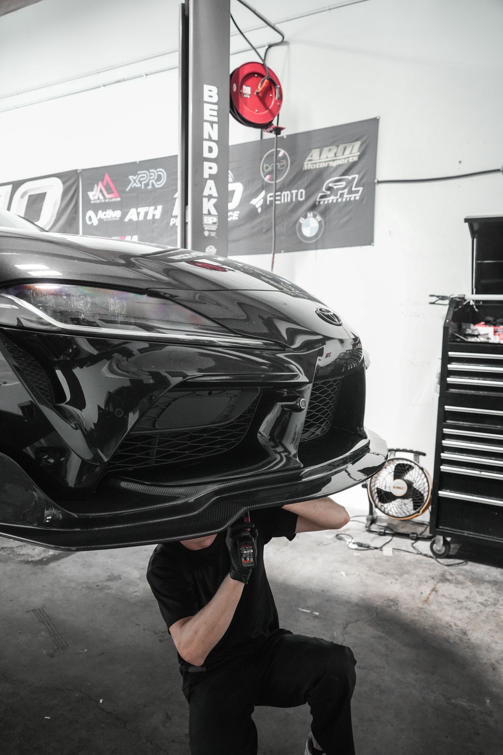 Mechanic crouching under a black car in an auto shop, with tools and tire racks nearby.