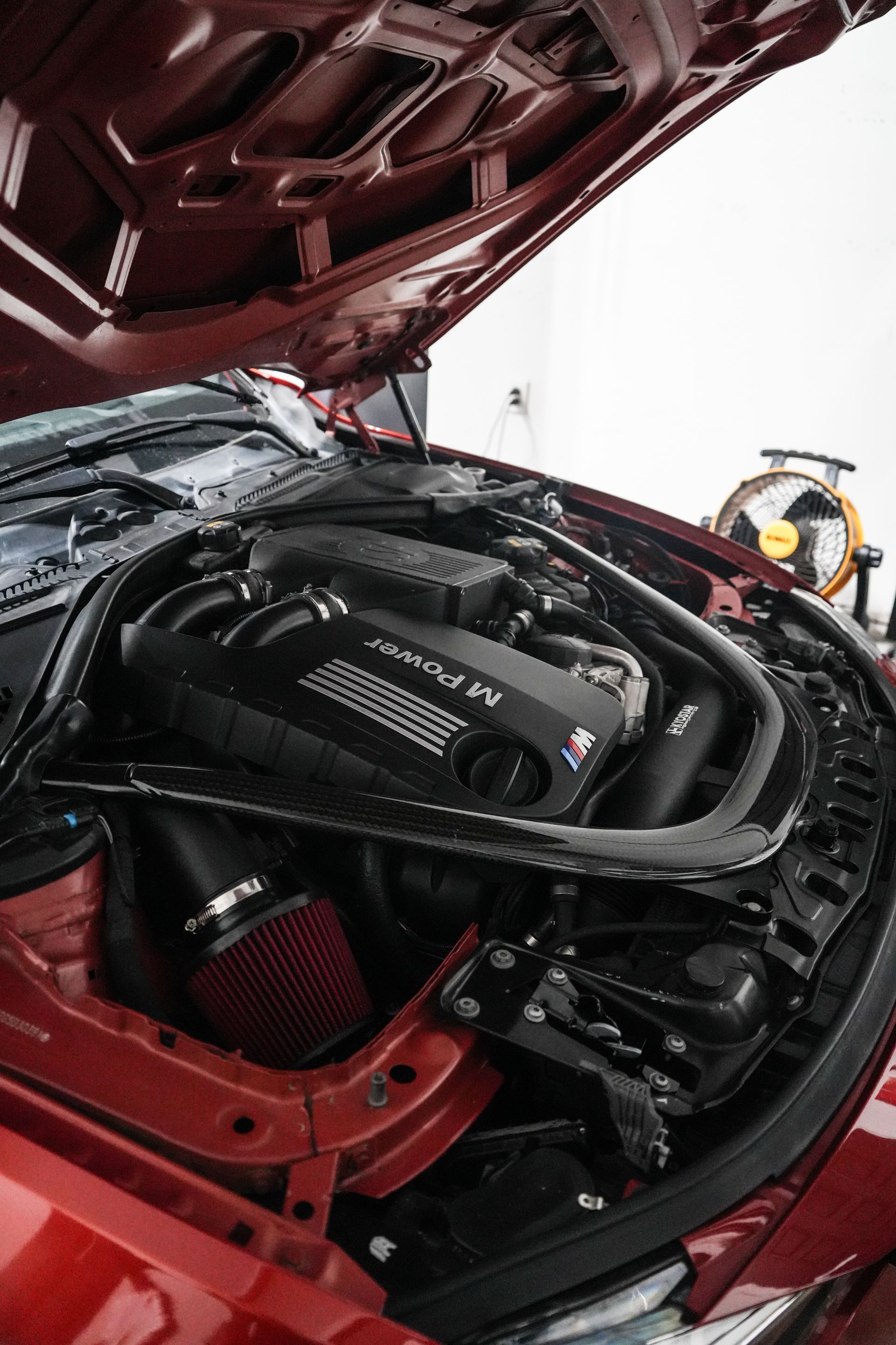 Open car hood showing a red vehicle’s engine bay with visible hoses and engine components