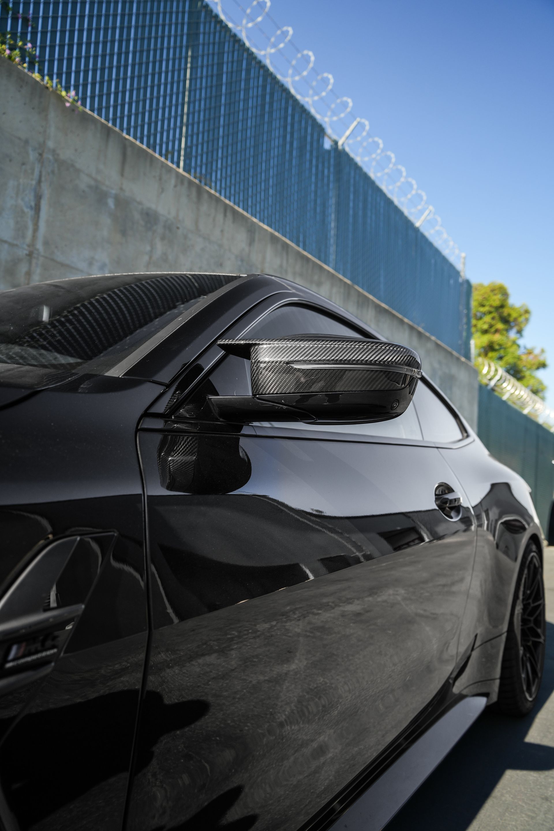 Black car parked beside a concrete wall topped with barbed wire under a blue sky