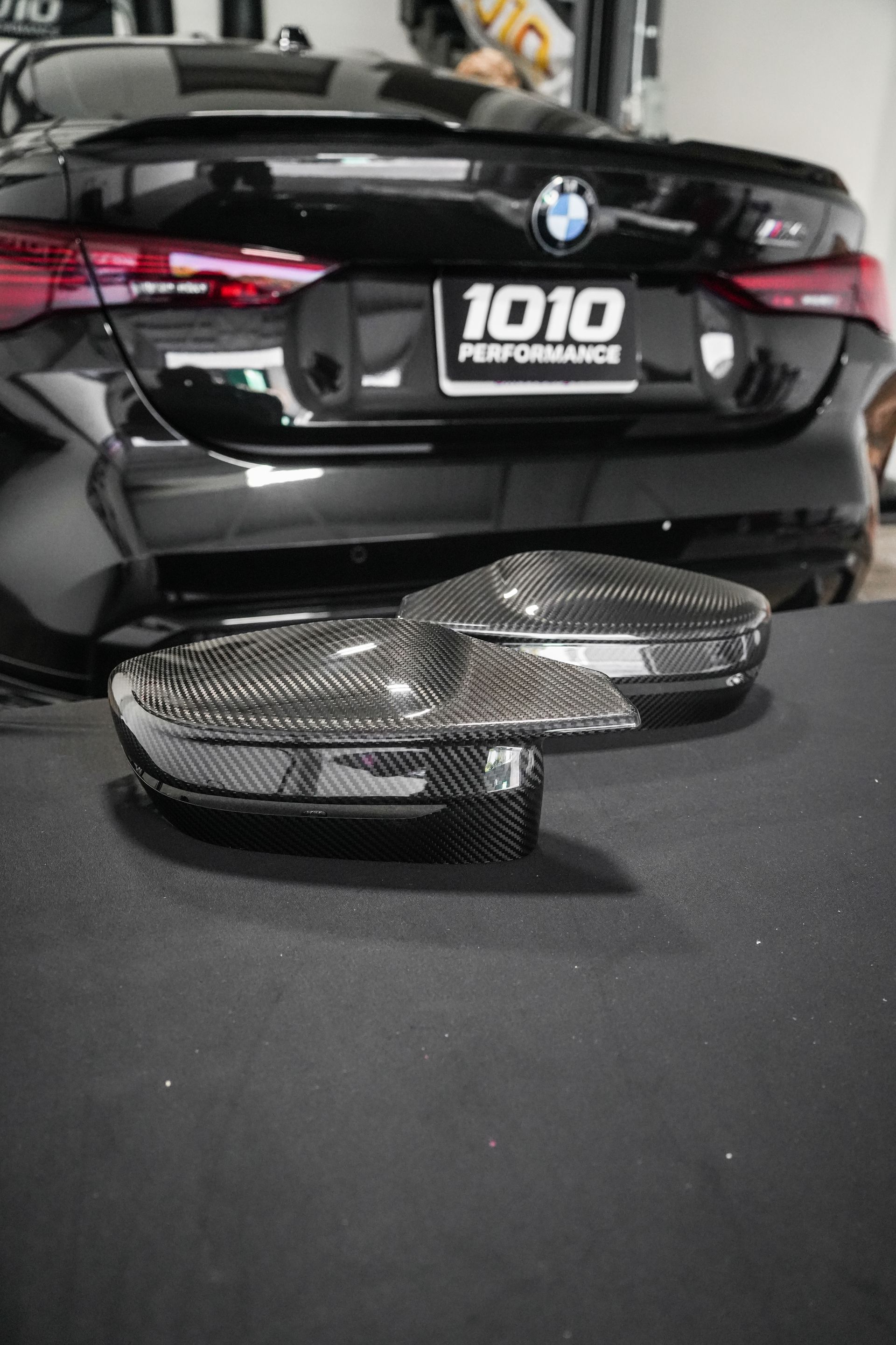 Two carbon fiber car side mirror caps on a black surface in front of a black BMW in a garage.