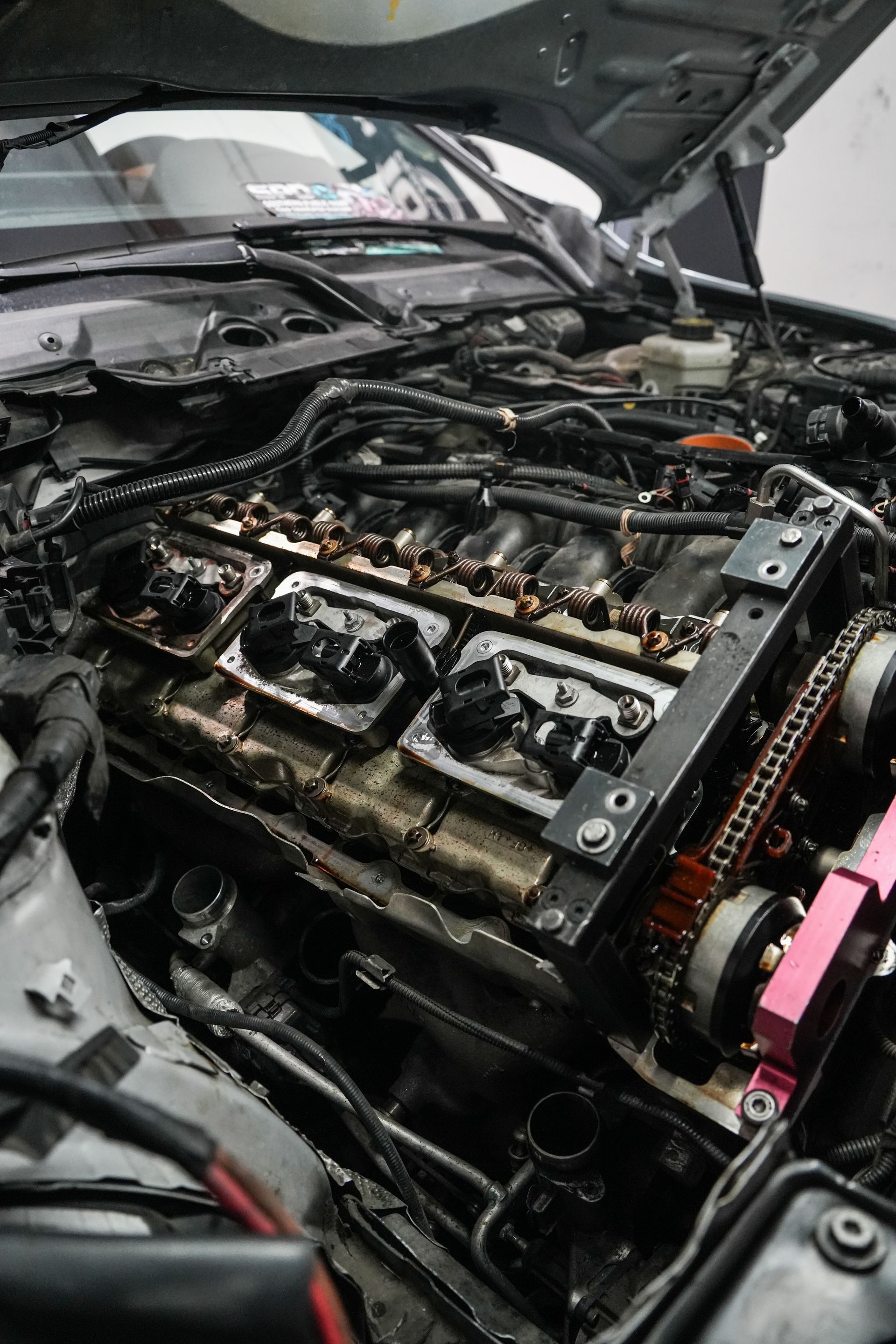 Exposed car engine under an open hood, with visible cylinder head and wiring in a workshop.