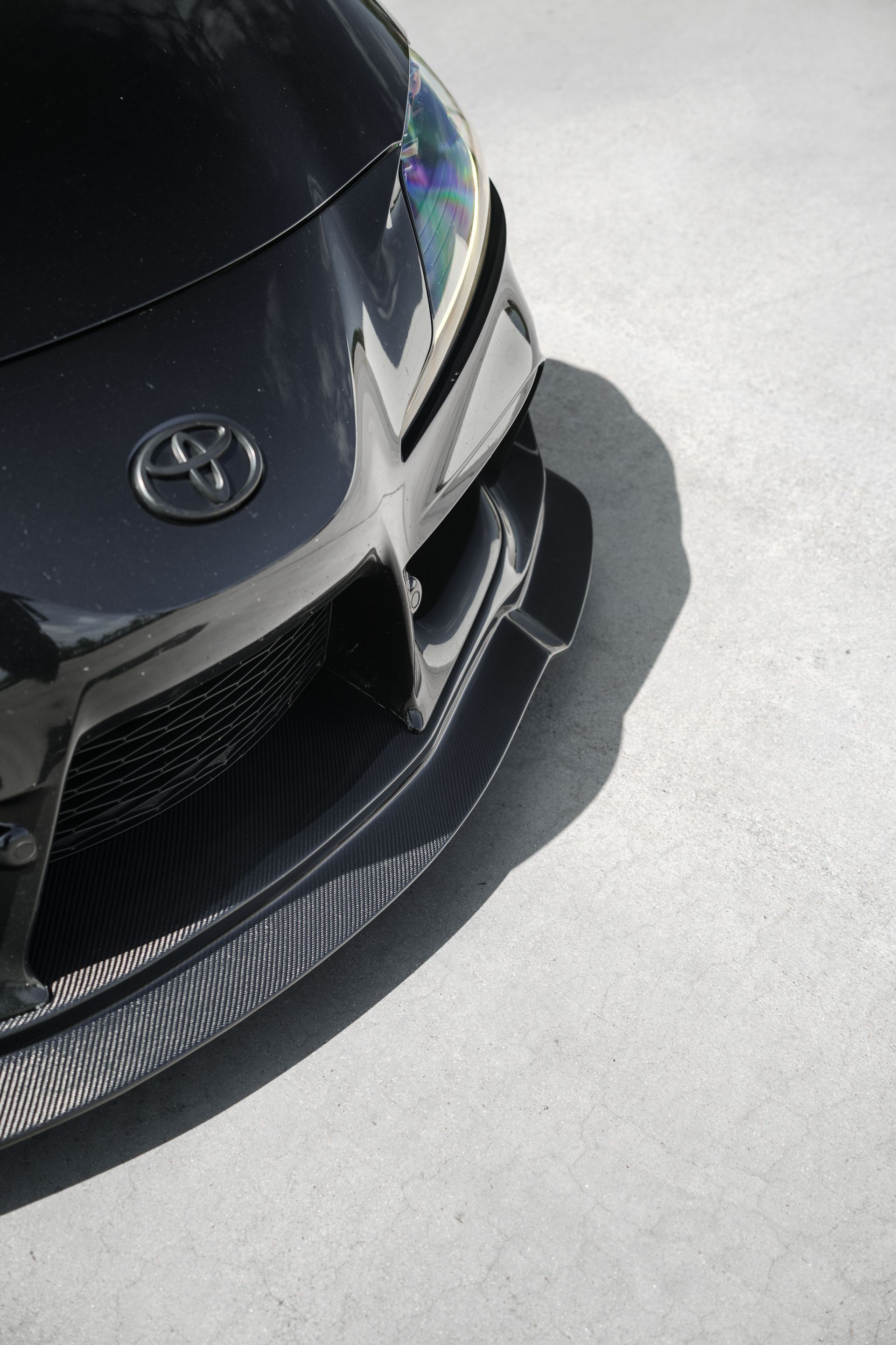 Front view of a black Toyota car with a carbon-fiber front splitter on a sunlit pavement.