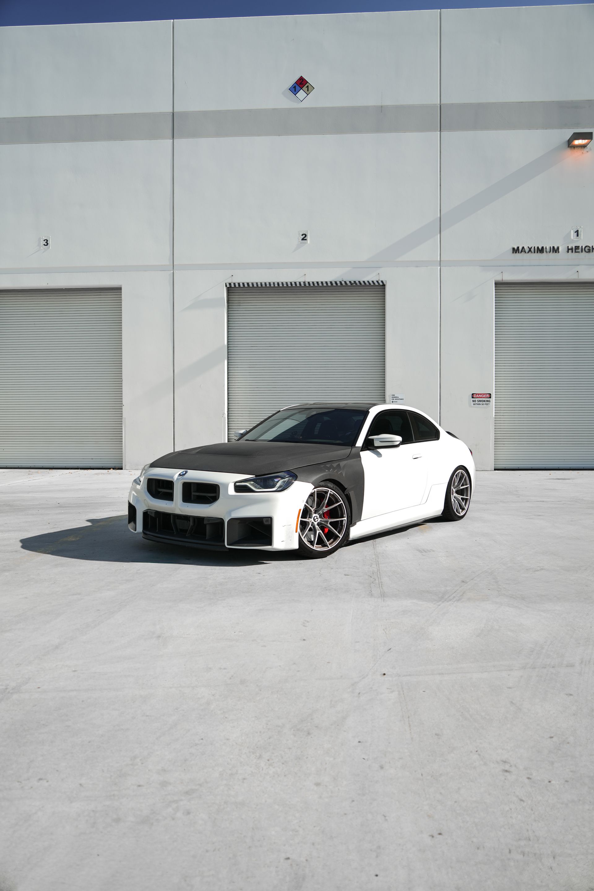 White sports car with exposed front body kit parked outside a gray industrial building.