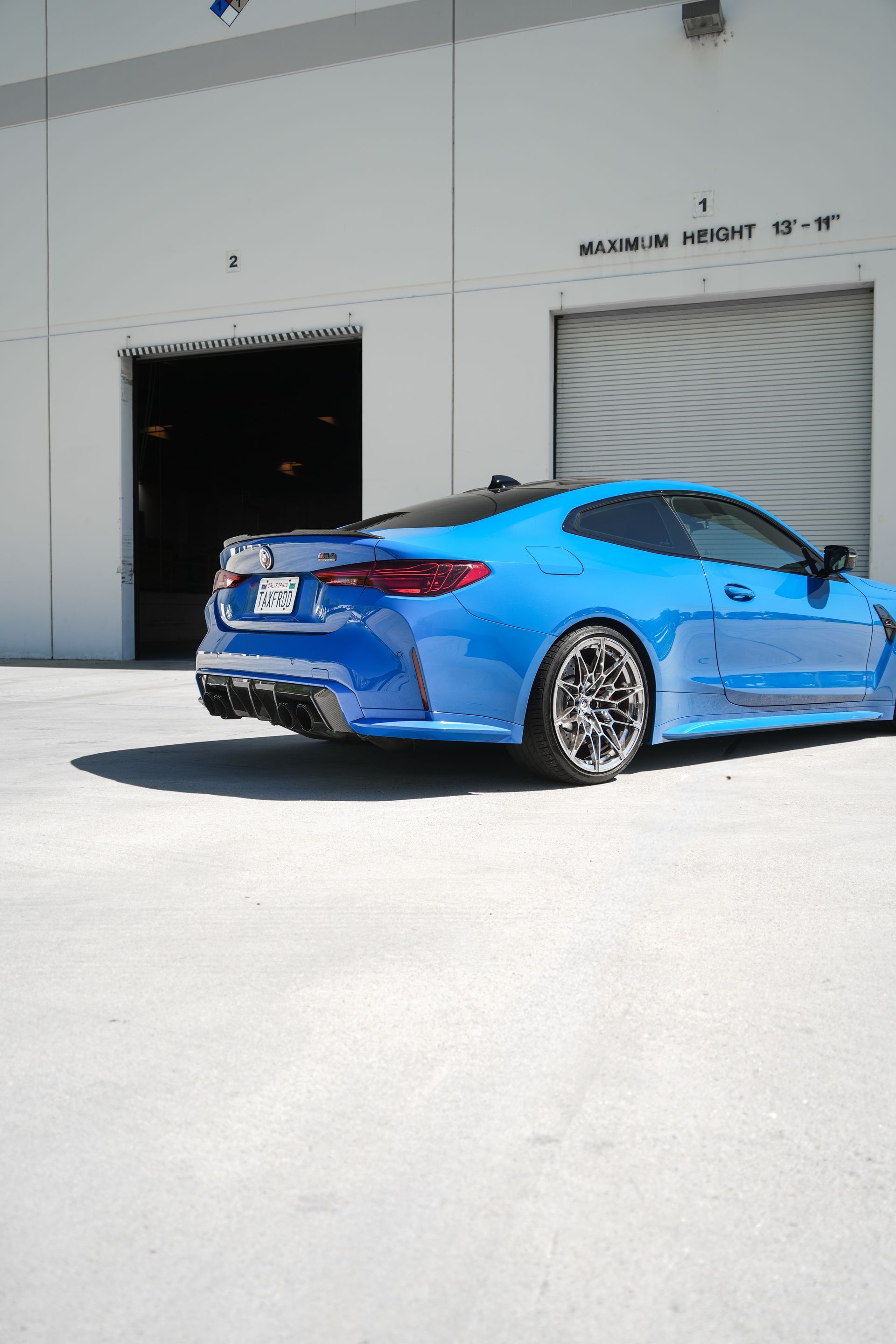 Blue sports car parked outside a warehouse with an open loading bay and closed roll-up door.