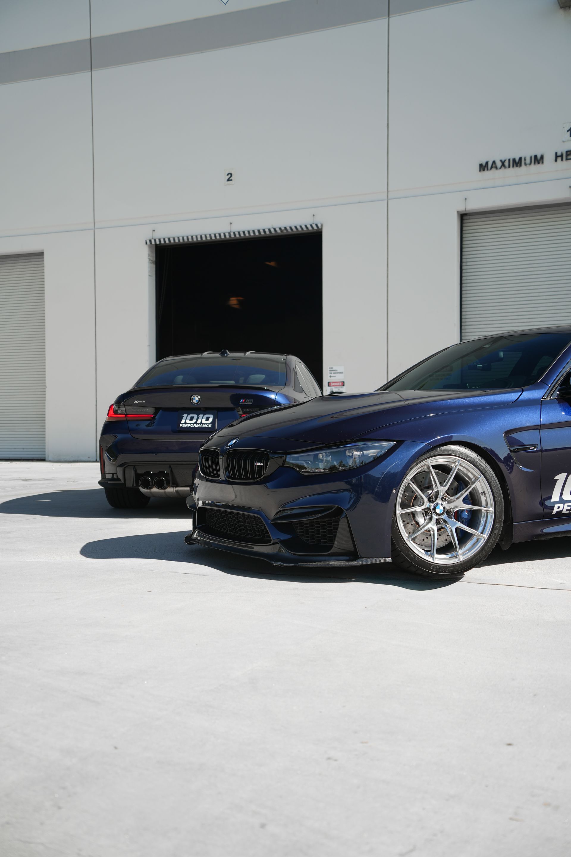 Two black BMW sedans parked outside a white garage, one in the foreground with silver wheels.