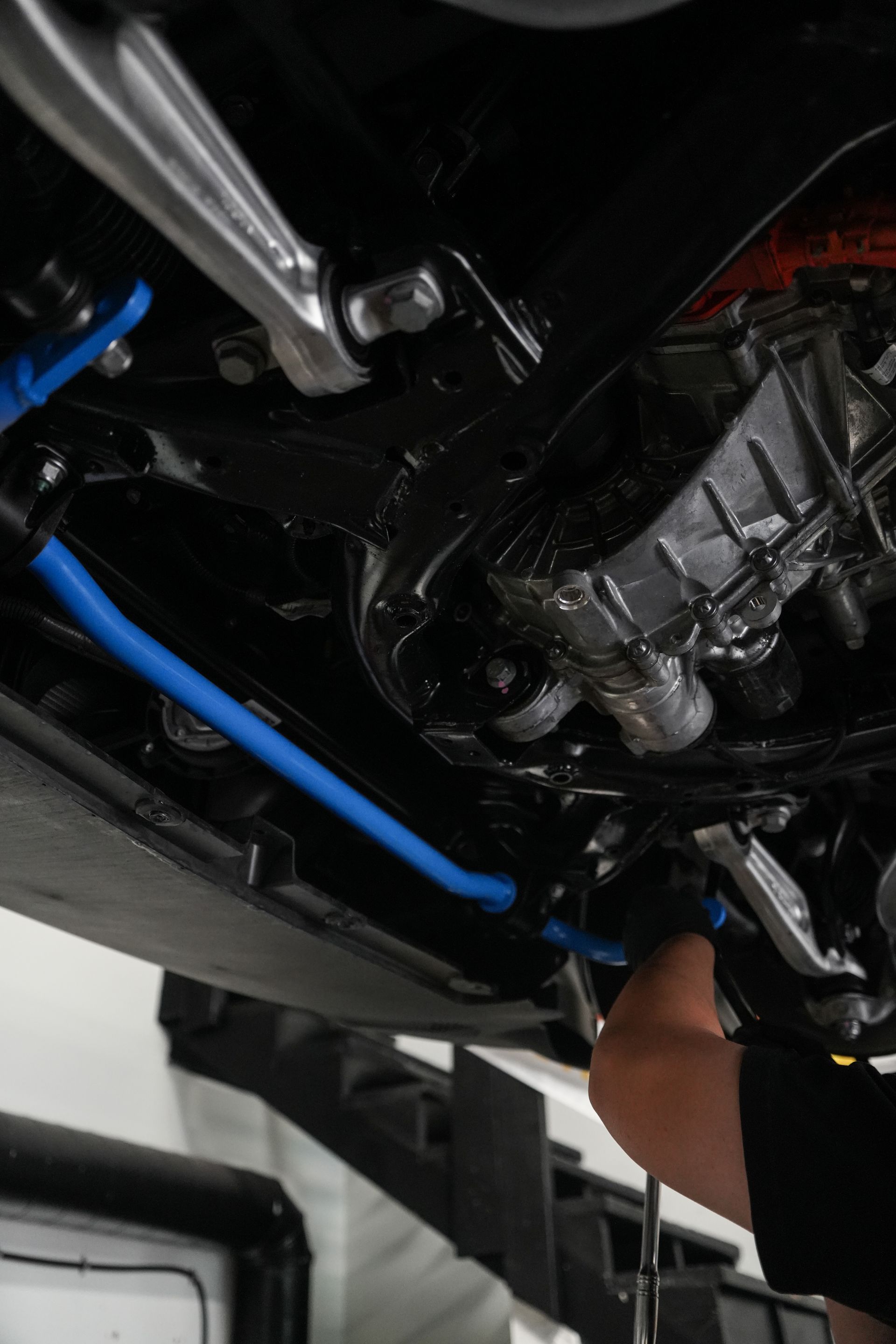 Underneath a car, a person works on the engine with blue hoses and metal tools visible.