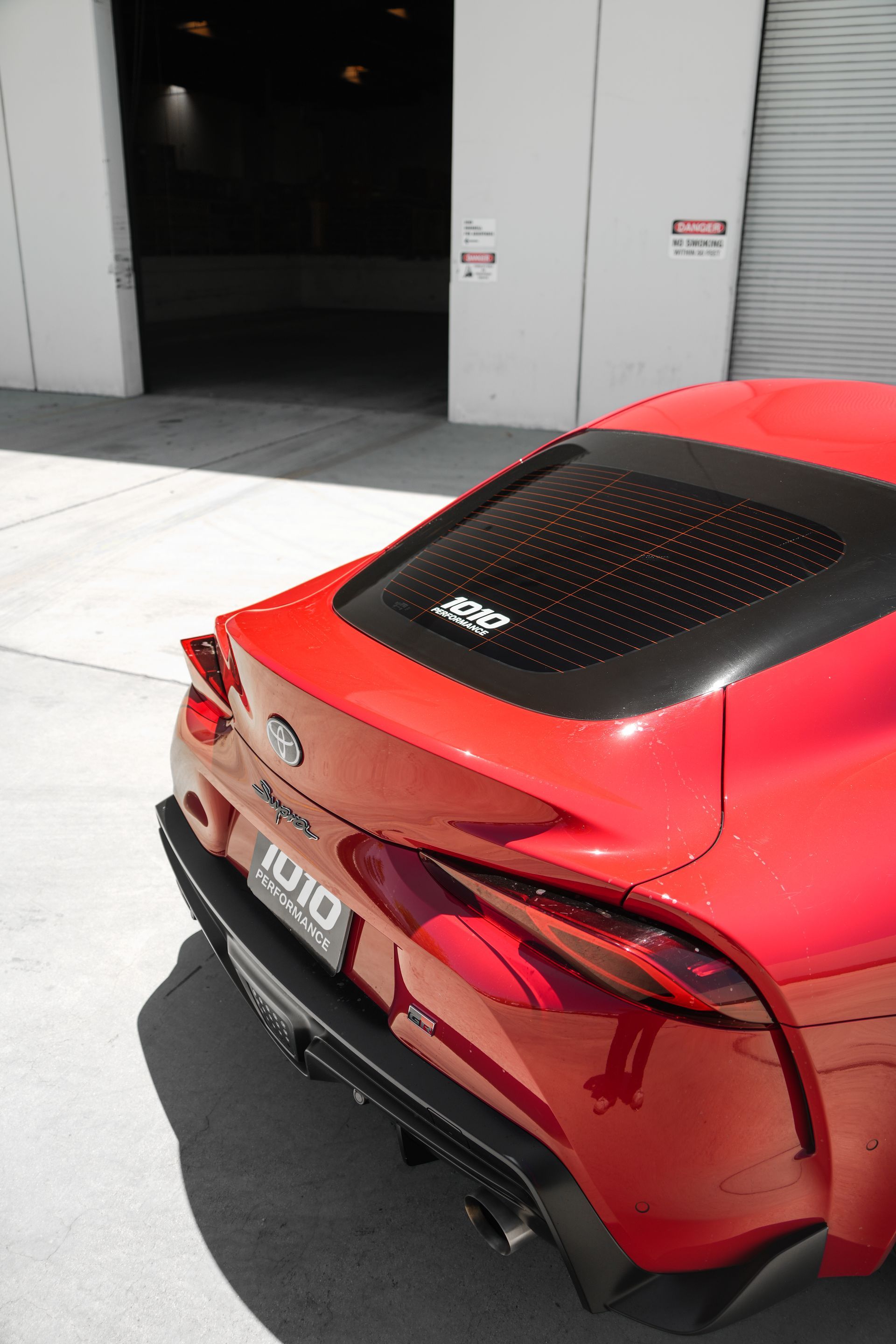 Red sports car parked outside a building, rear view with black spoiler and roof panel.
