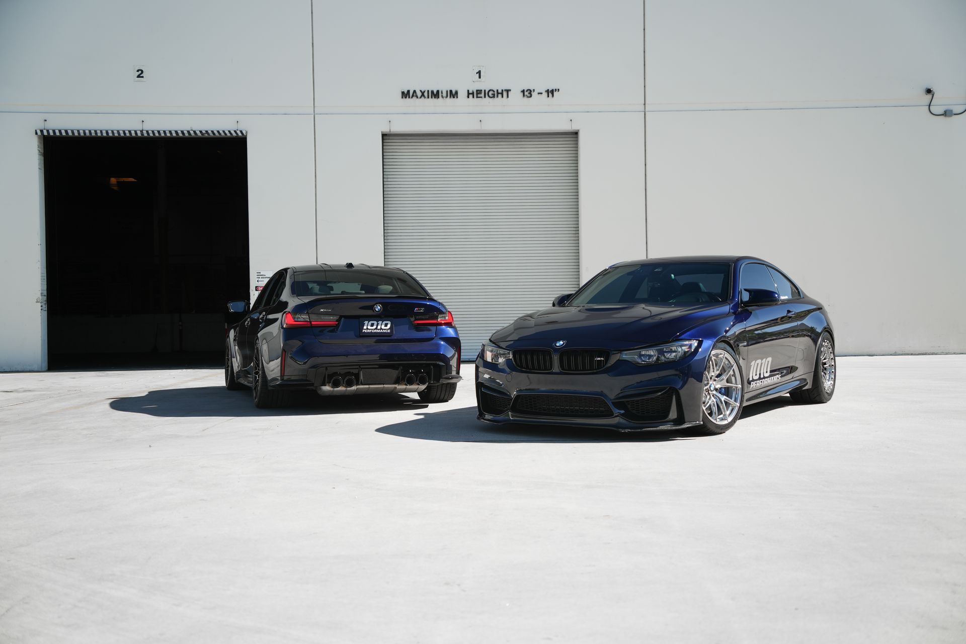 Two black cars parked outside a white industrial building, one in front and one near a garage door