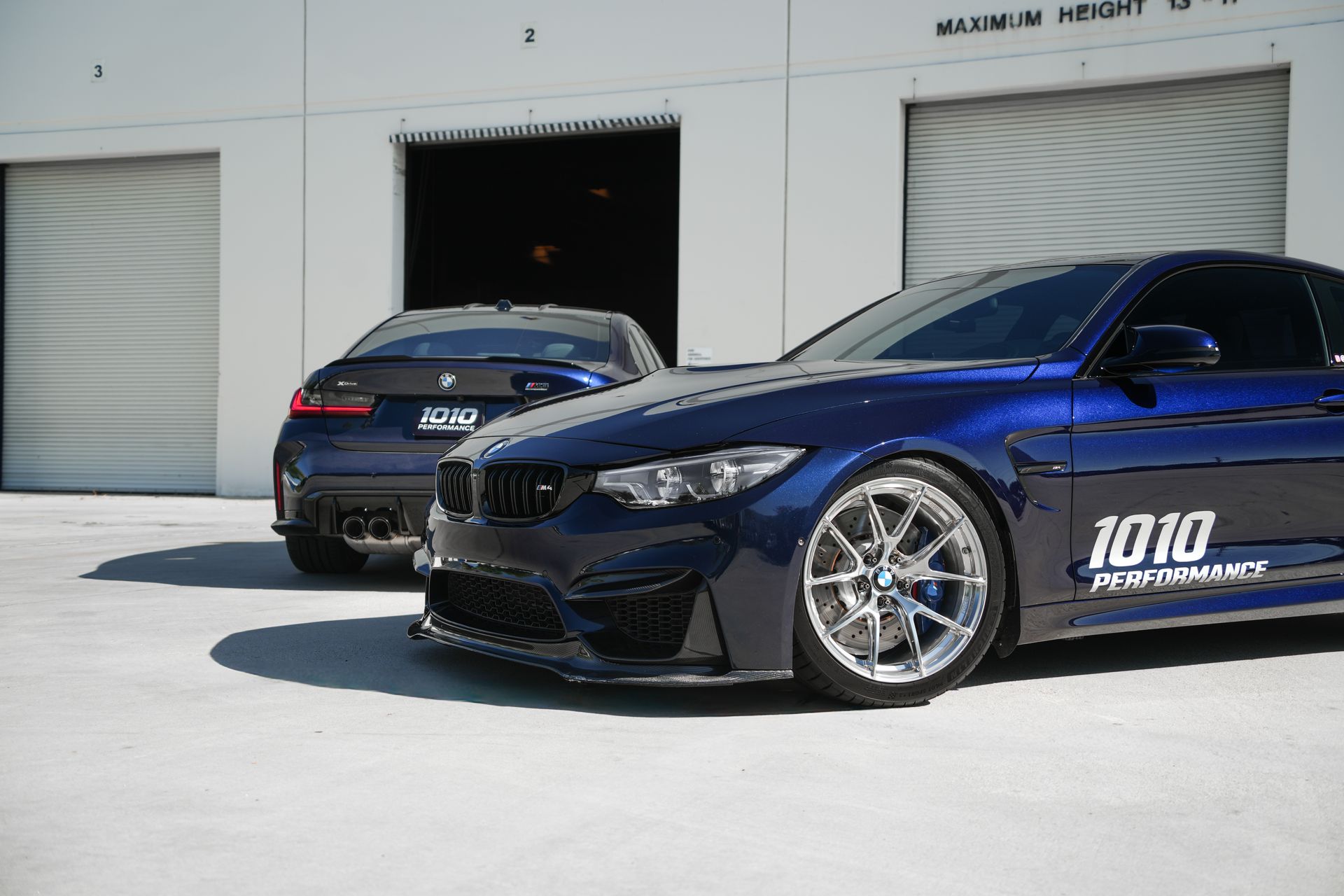 Two blue BMWs parked outside a garage, one with a black hood and racing decals.