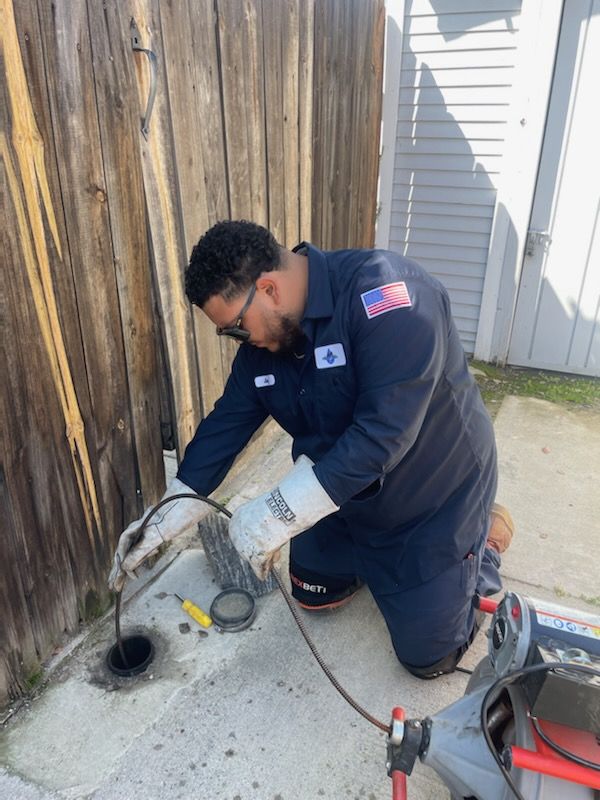 Plumber in blue uniform kneels, using a drain snake on a drain outside.  Sunlit area next to wooden fence.