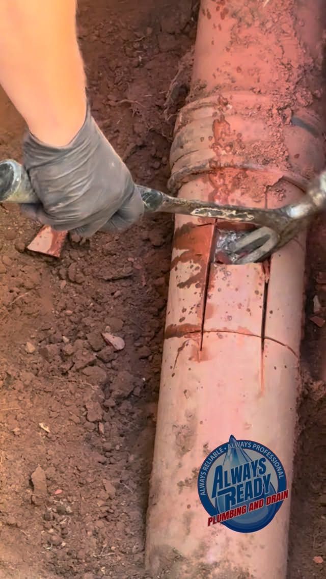 A gloved hand uses a wrench to cut a damaged orange pipe in a dirt trench.