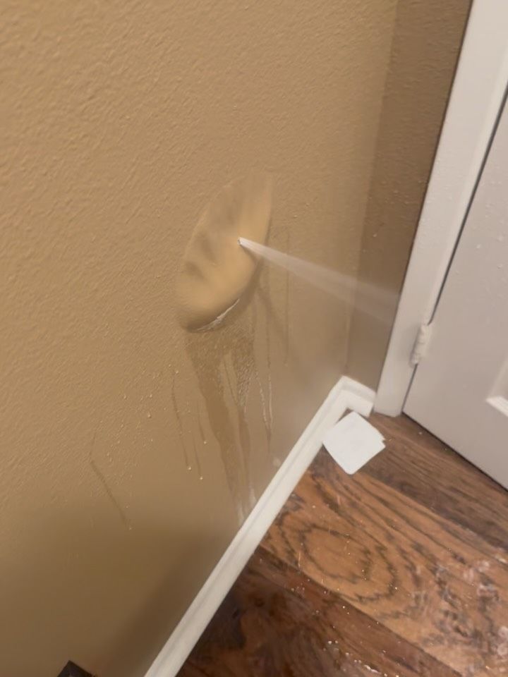 Water spraying from a hole in a beige wall, damaging the wall and floor.