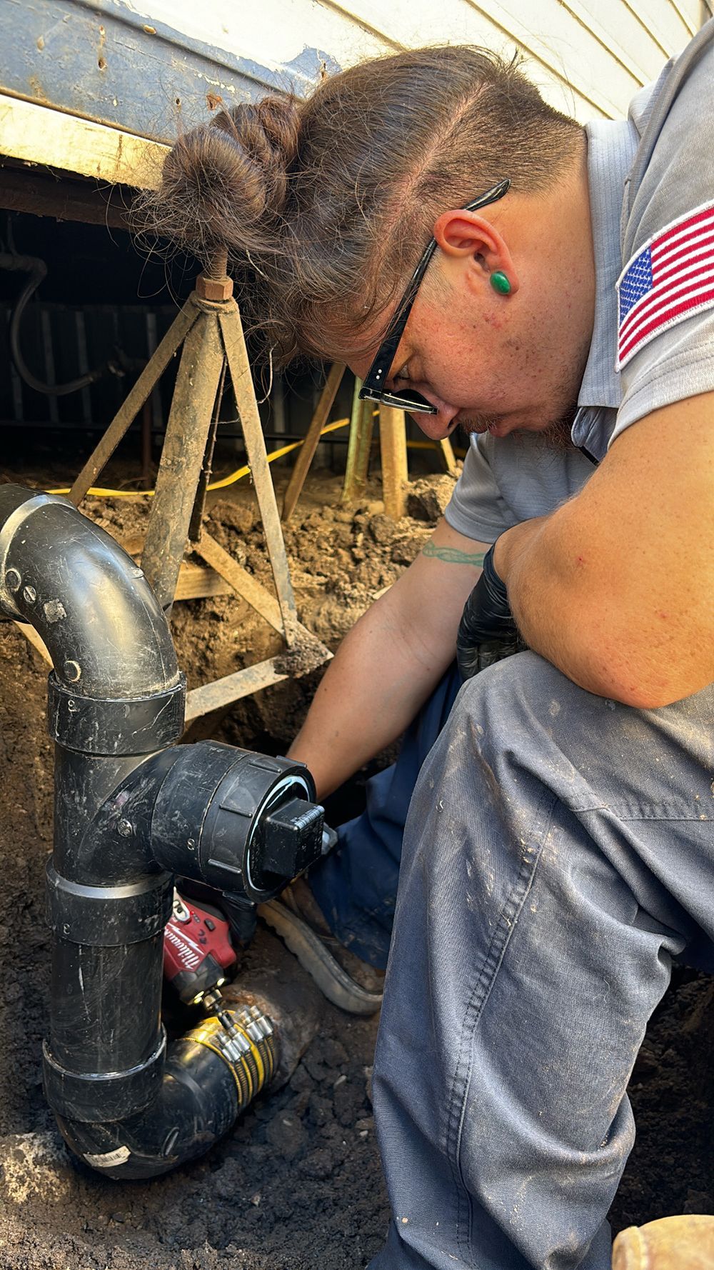 A person in work clothes repairing a black pipe in a dirt-filled trench.