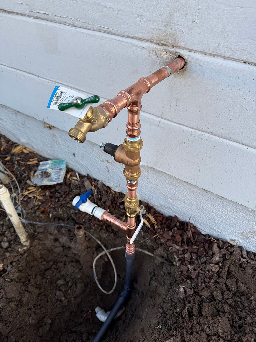 Copper pipes and valves installed on the exterior of a white building for water supply, near a dug hole.