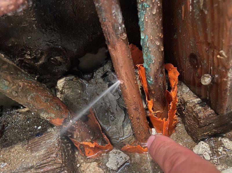 Leaking copper pipes with corrosion and a finger pointing to the leak, in a damp, enclosed setting.