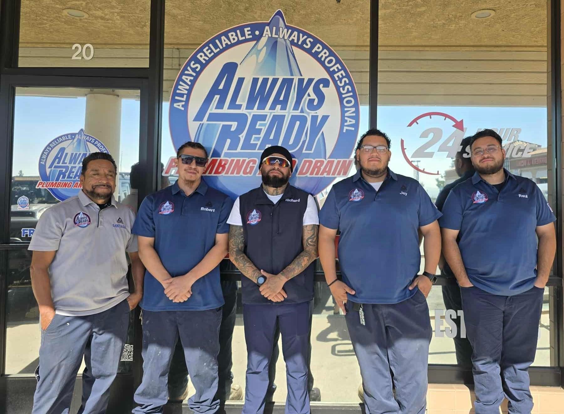 Five people in blue work uniforms stand outside a plumbing business with a logo reading 