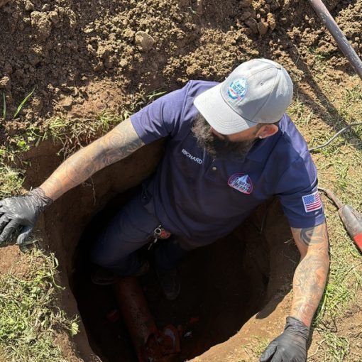 Plumber in a deep hole, inspecting a pipe. He wears a hat, gloves, and is surrounded by dirt.