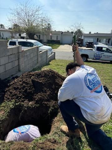 Two workers in white shirts dig in a yard, one in the hole. Houses and a truck are in the background.