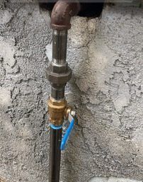Black pipe with a brass valve and blue handle against a textured gray wall.