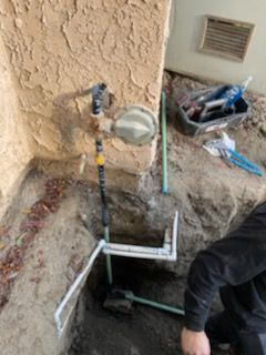 Plumbing work in progress: pipes, valve, and a man in a black jacket digging near a stucco wall.