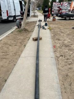 A long, black pole lies on a sidewalk, with workers and vehicles visible in the background.