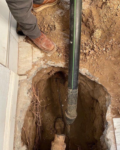 Plumber working on a pipe in a hole in the floor; black pipe, dirt, and brown boots are visible.
