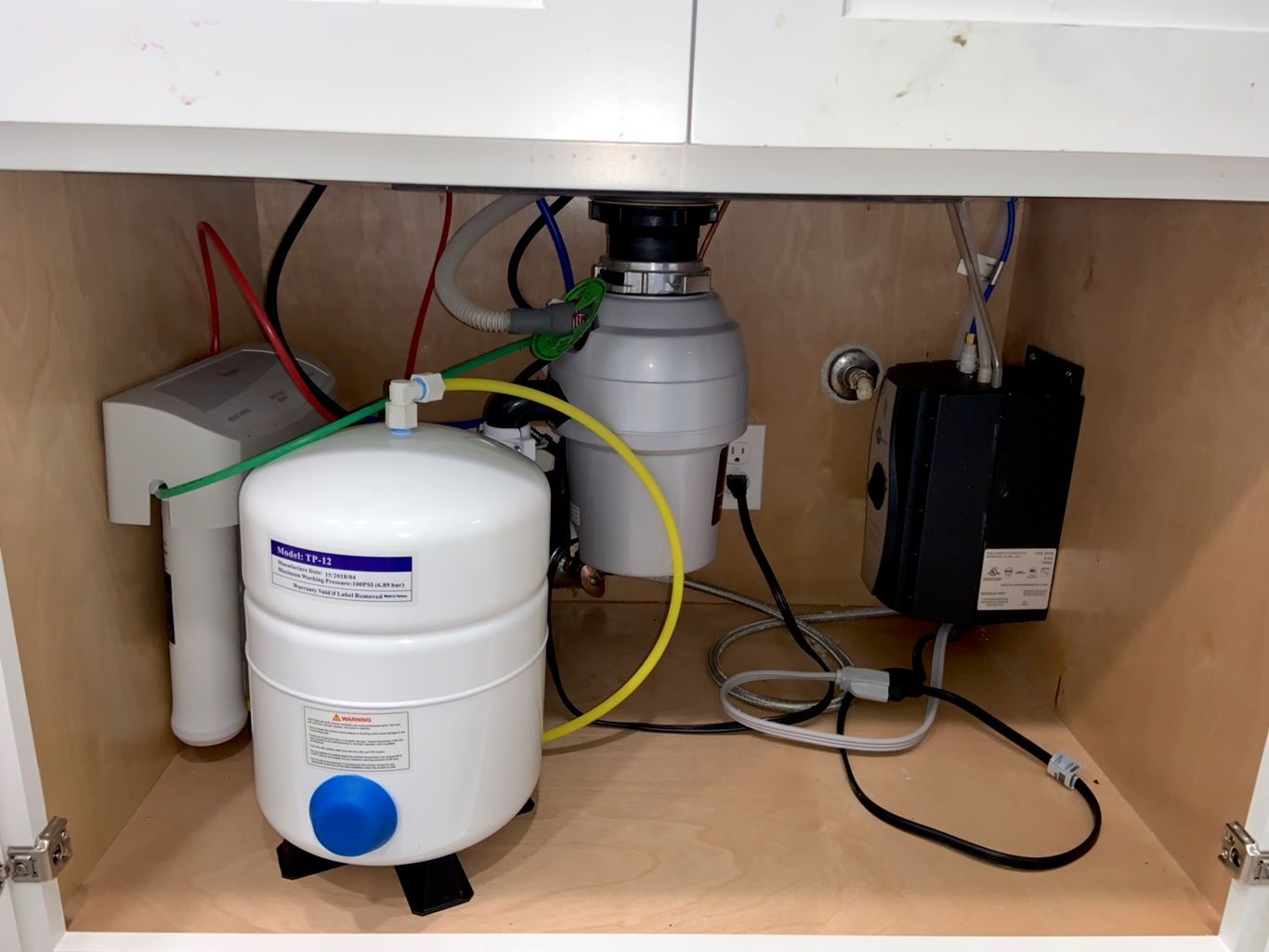Under-sink view of white water filter, garbage disposal, and black electrical box with various tubes and wires.