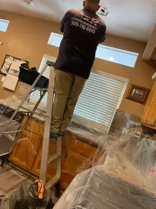 Man on a ladder reaching towards a ceiling fixture in a kitchen with covered cabinets.