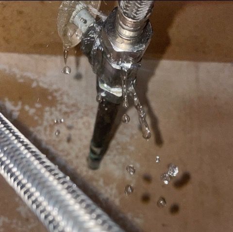Water leaking from a metal pipe and braided hose connection in a brown setting. Water droplets are visible.