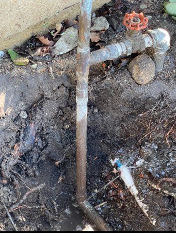 Exposed outdoor water pipe, valve, and fittings in muddy ground, near a concrete wall.