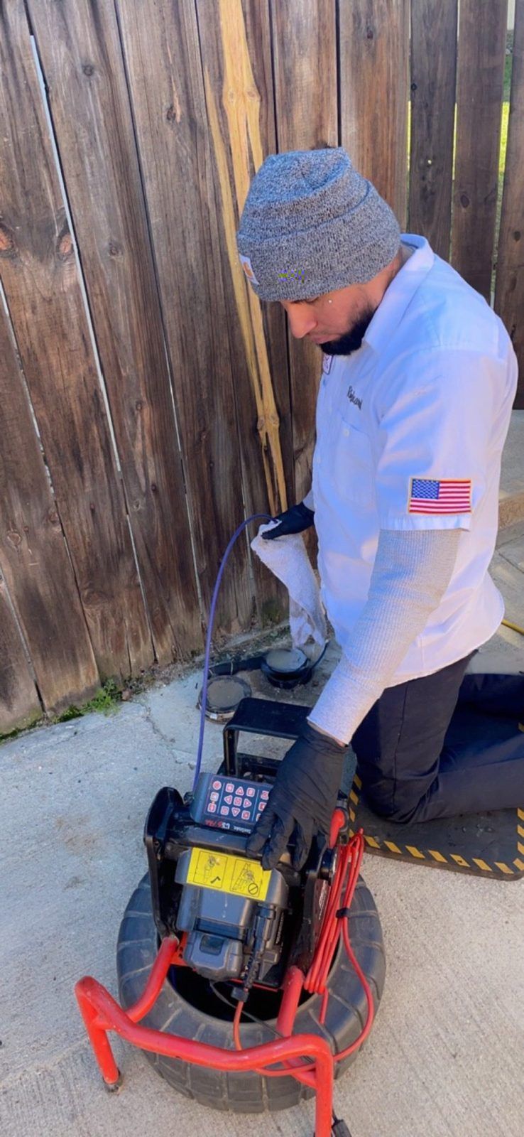 A person using a drain camera outside. They wear a beanie, work gloves, and a white shirt with an American flag patch.