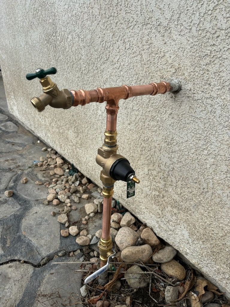 Outdoor water faucet with copper pipes and a backflow preventer against a stucco wall.