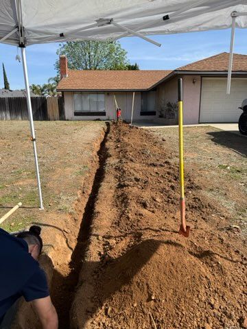 A man digging a trench in a yard under a canopy. A house is in the background.