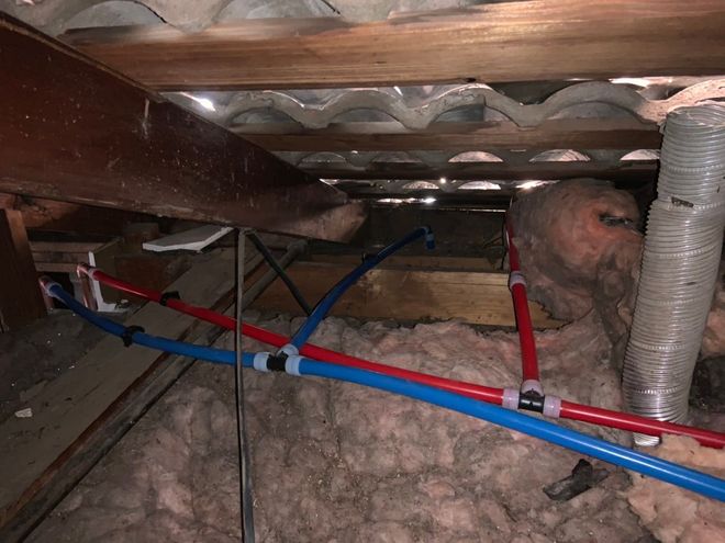 Pipes in an attic. Blue and red PEX plumbing against insulation. Wooden beams and corrugated roofing overhead.