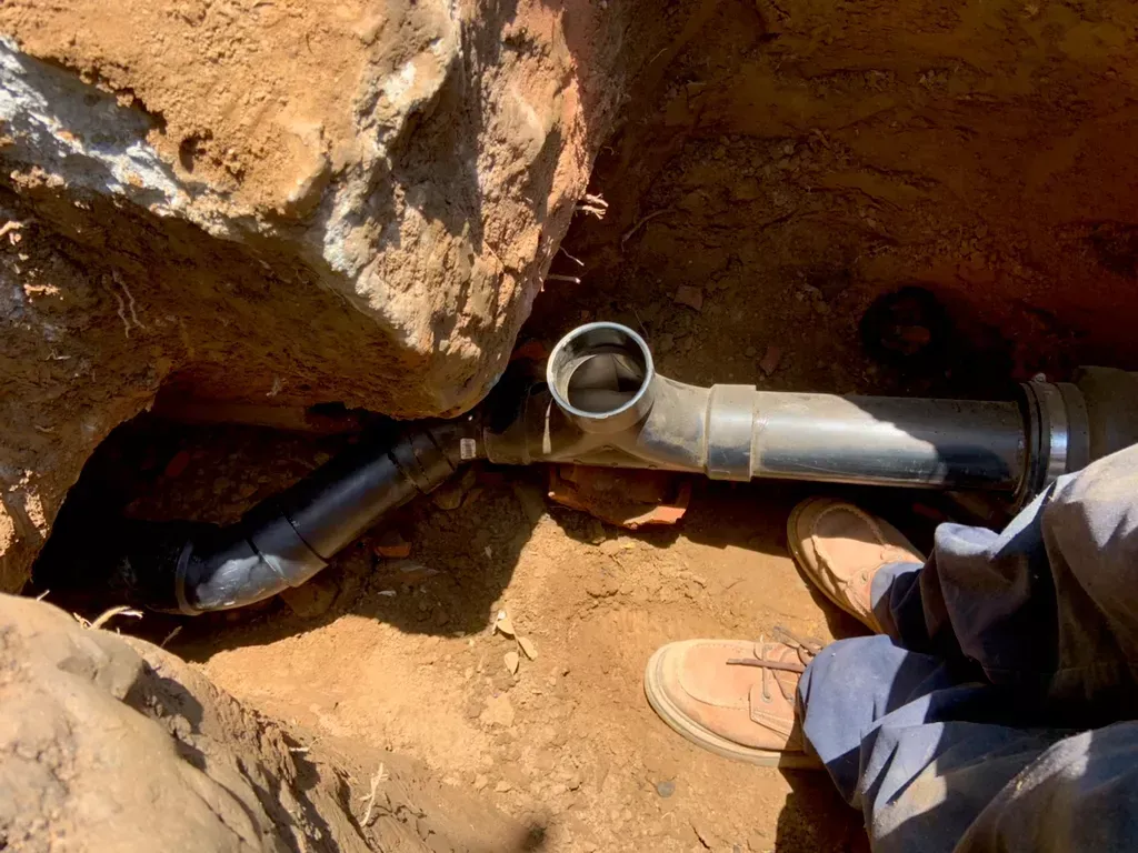 Black plumbing pipes and fittings in a dirt trench, with person's legs and work boots visible.