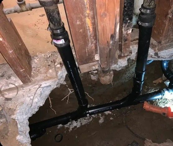 Black plumbing pipes in a concrete hole, connected to older pipes and a wooden wall.