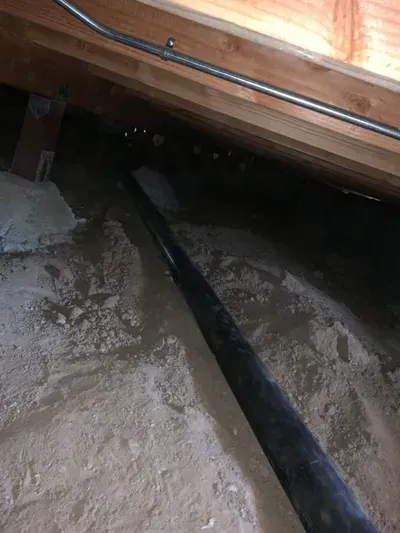 Underneath wooden framing, a black pipe extends across a concrete-like surface in a dark, confined space.