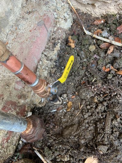 Copper pipes connected to a valve with a yellow handle, surrounded by dirt and concrete.