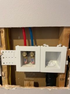 Washing machine hookup in a wall: water valves (red/blue pipes) and drain in a white box.